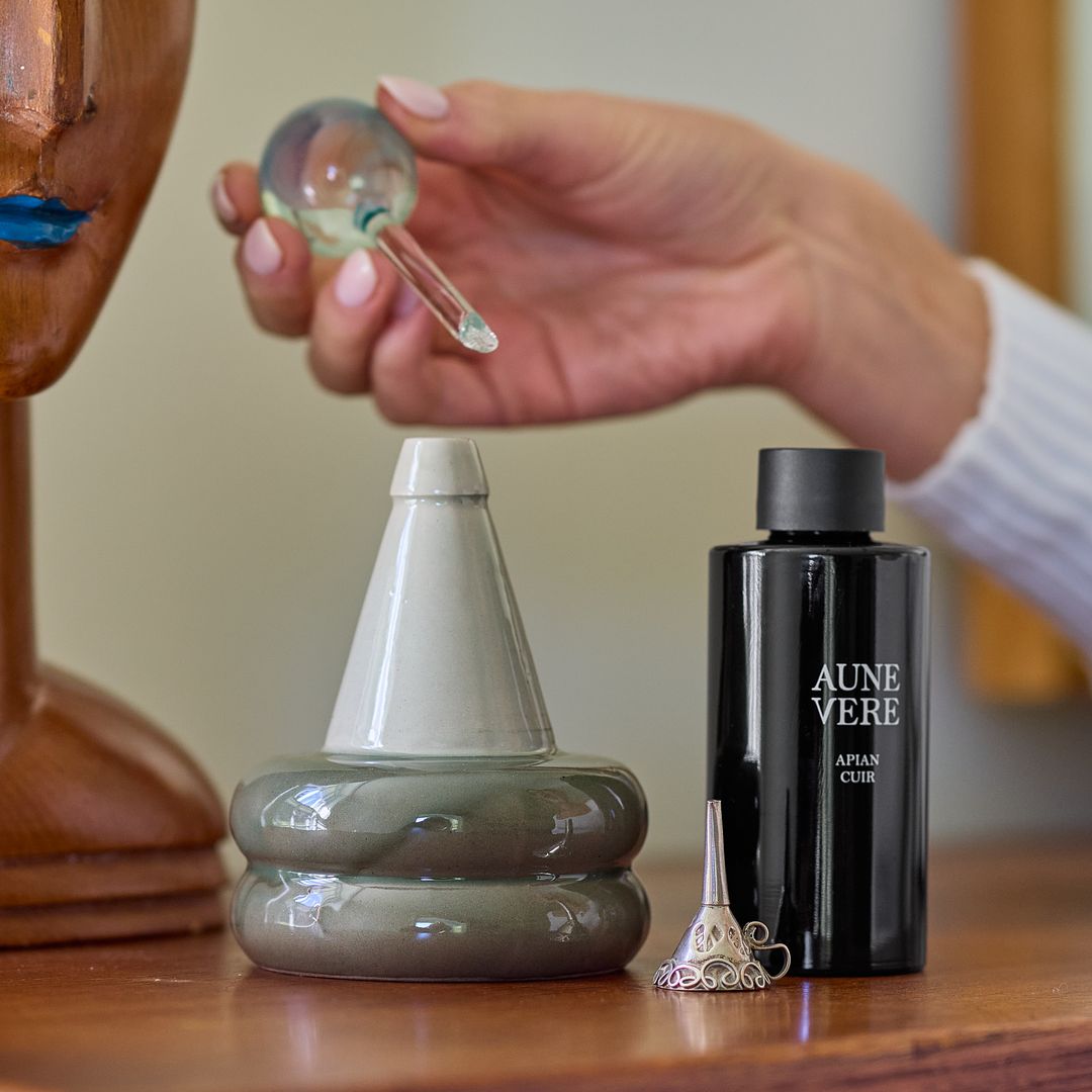 A hand holds the Apian Cuir applicator above the open ceramic vessel, the 100 ml bottle standing beside it.