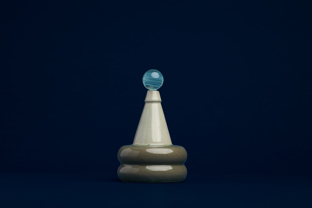Apian Cuir ceramic vessel, a stacked glazed form holding a glass orb, on deep navy paper.
