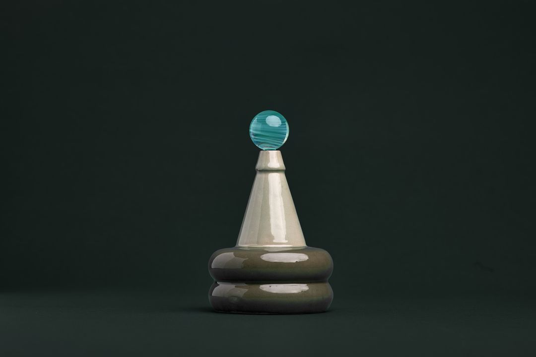 Apian Cuir ceramic vessel, a faceted dark glazed base holding a clear glass orb, on gray green paper.