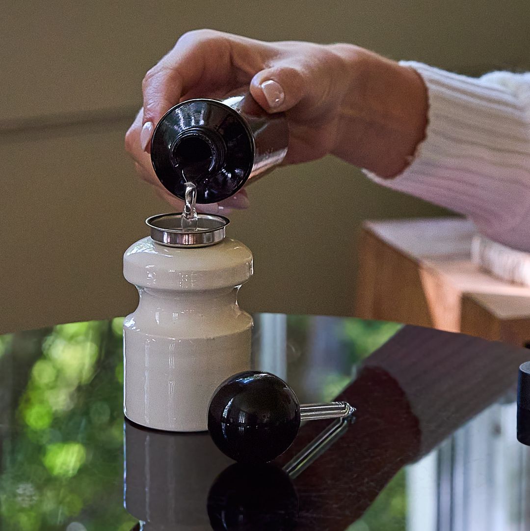 A hand pours Caledonian Pear from the 100 ml bottle into a pale ceramic vessel, the applicator resting on a glass table.