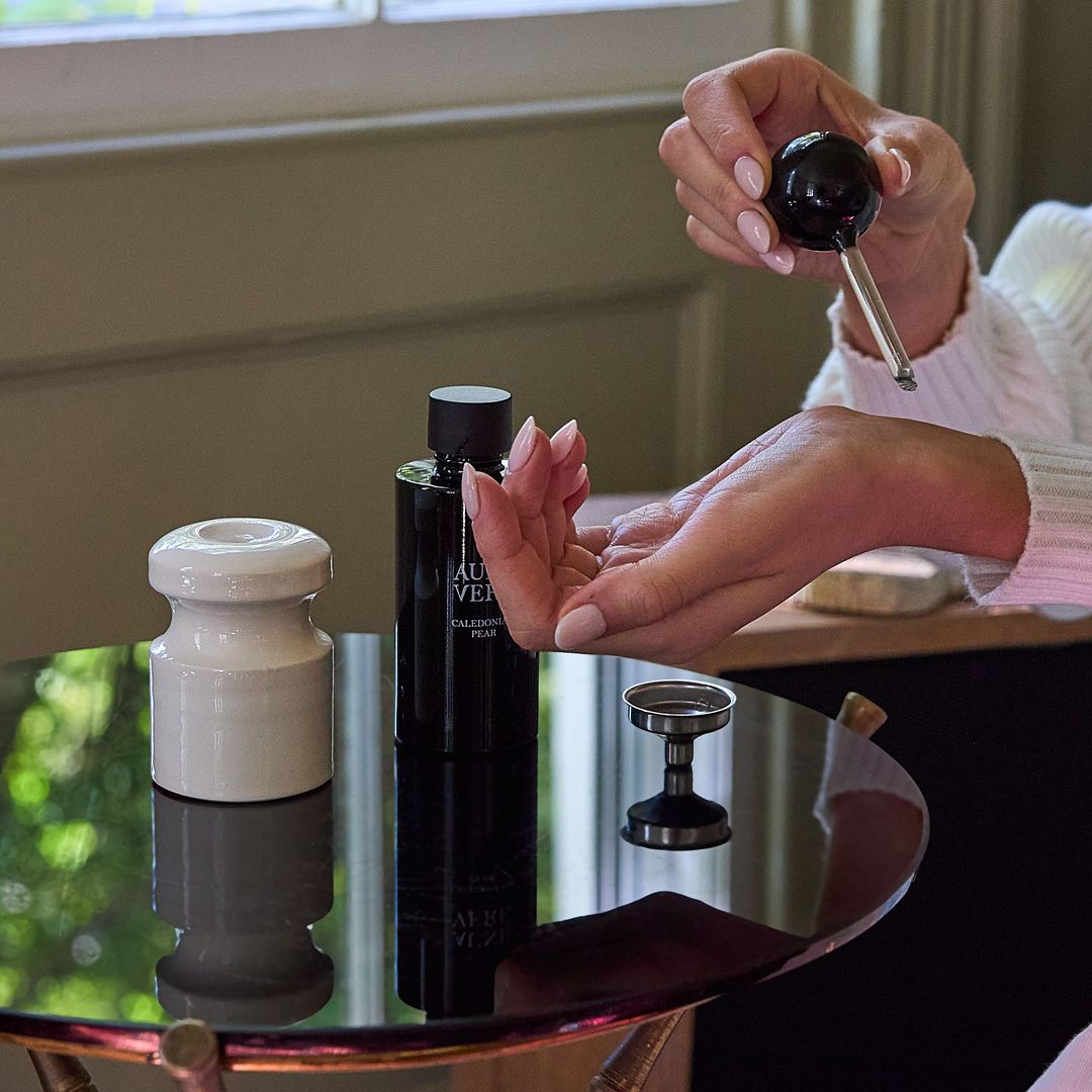 A hand holds the Caledonian Pear applicator above an open palm, the 100 ml bottle and a pale ceramic vessel on a glass table.