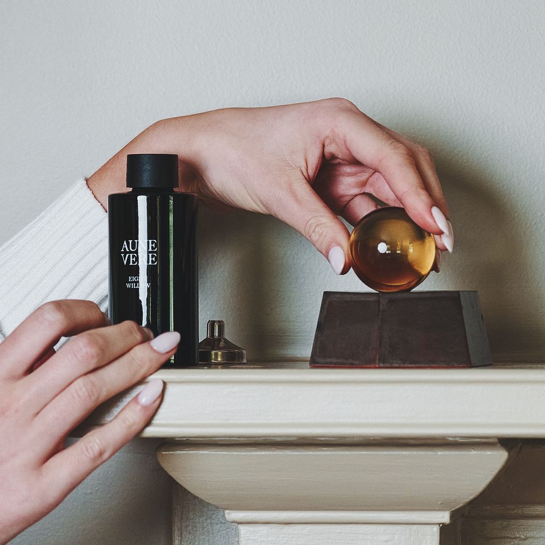 Hands set the amber glass orb onto the Eight Willow vessel on a mantel, the 100 ml bottle standing beside it.