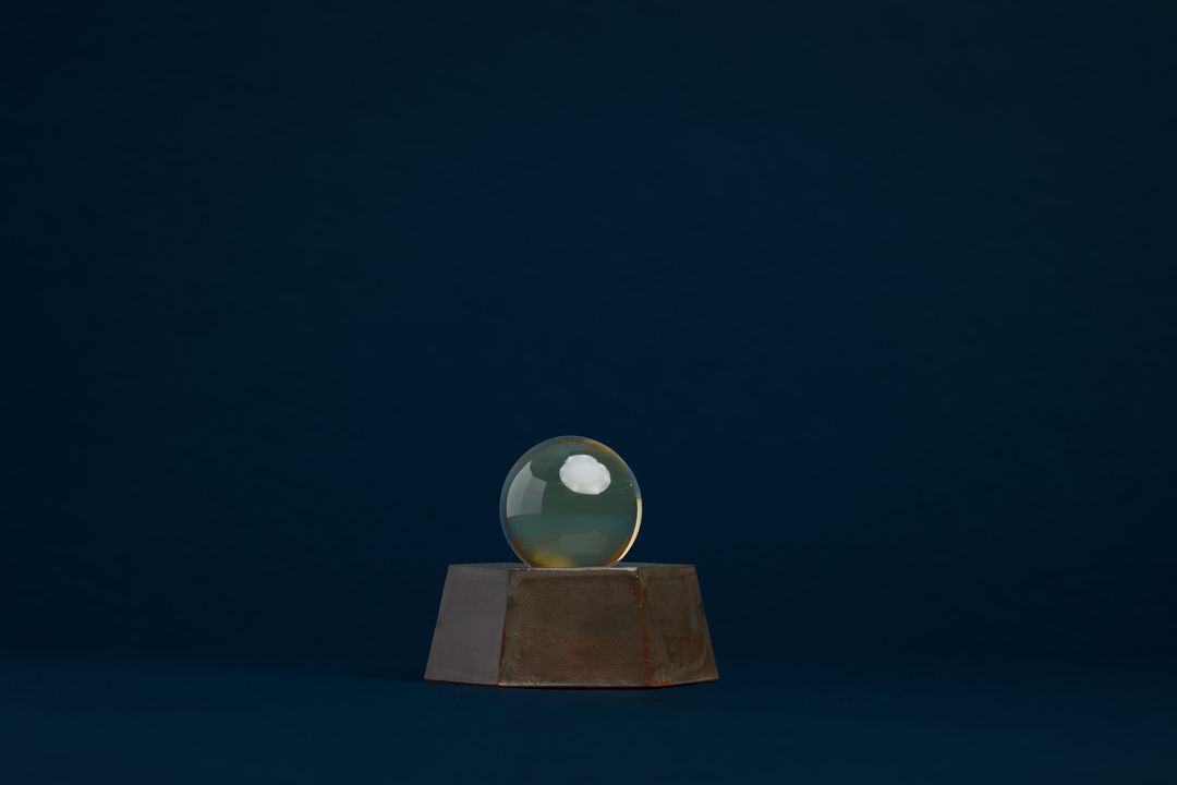 Eight Willow ceramic vessel, a faceted dark glazed base holding a clear glass orb, on deep navy paper.