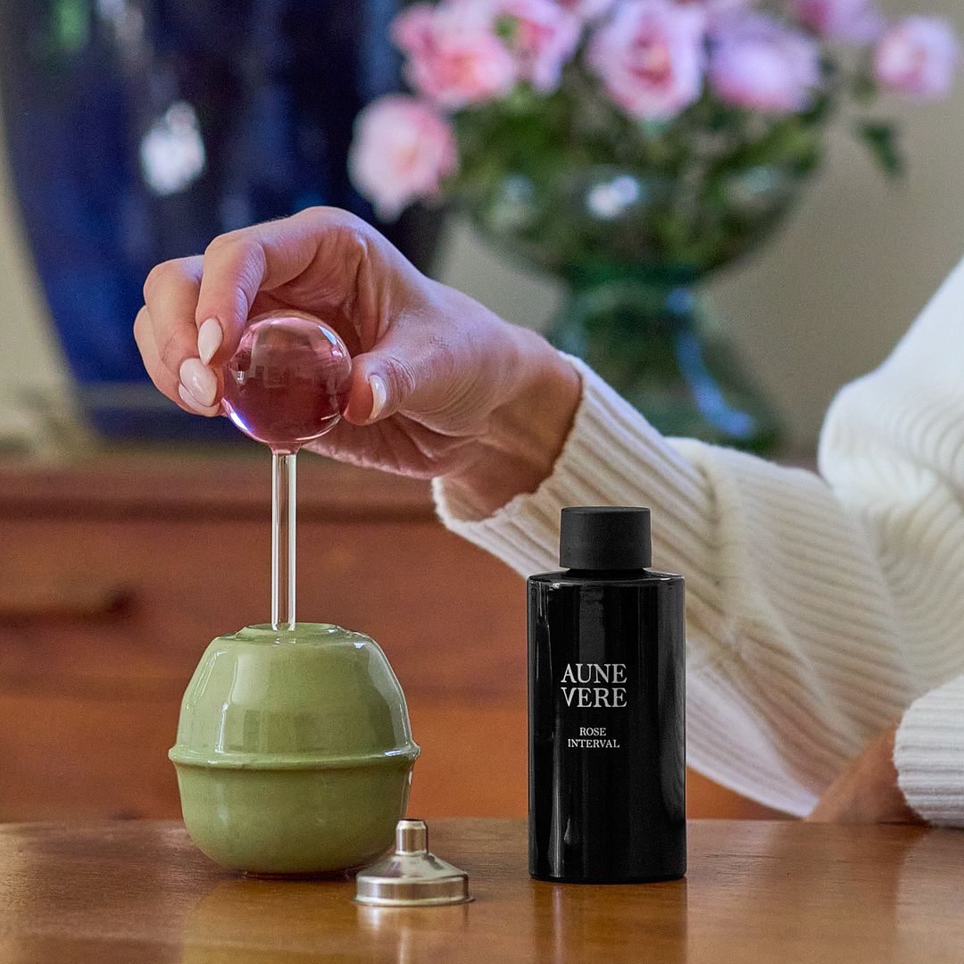 A hand lifts the pink glass applicator from the Rose Interval vessel, the 100 ml bottle standing beside it on a wooden table.