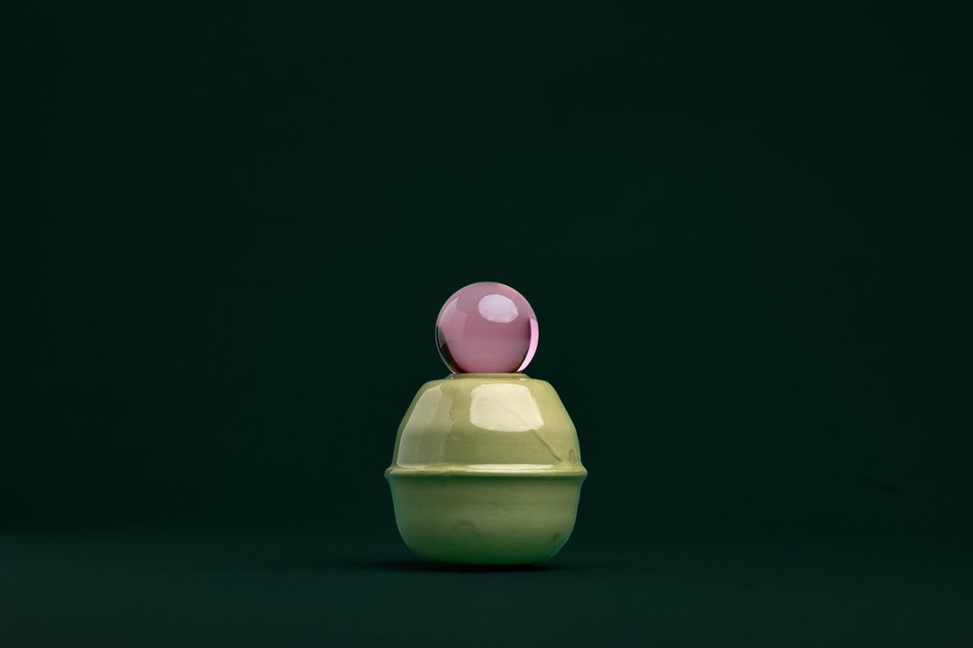 Rose Interval ceramic vessel, a faceted dark glazed base holding a clear glass orb, on deep bottle green paper.