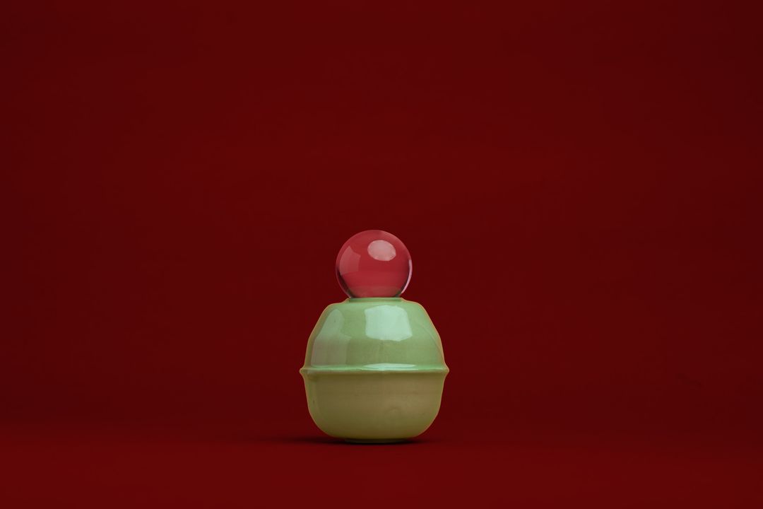 Rose Interval ceramic vessel, a pale green glazed form holding a glass orb, on alder red paper.